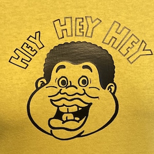 May include: A yellow t-shirt featuring a black cartoon illustration of a smiling, chubby person with the text "Hey Hey" at the top and bottom.