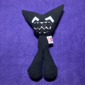 May include: A black plush toy featuring a wide, pointed head with two white triangular eyes and a large, jagged white mouth filled with black teeth. The body is slender with two long legs, and a small white tag is attached to the side.