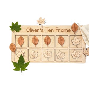 May include: A rectangular wooden ten frame board with the text Oliver's Ten Frame engraved on top. The board features ten square slots containing various wooden leaf-shaped tokens. Additional loose wooden leaf tokens and two green leaves are placed around the board, accompanied by a small white fabric drawstring storage bag.