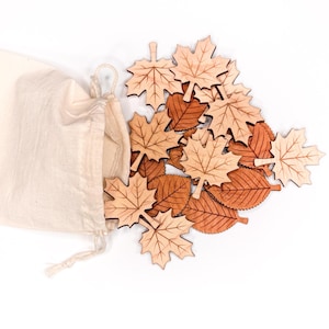 May include: A collection of laser-cut wooden leaf shapes in light and medium brown tones, featuring maple and rounded leaf designs with etched vein details, spilling out from an open, off-white cotton drawstring storage pouch.