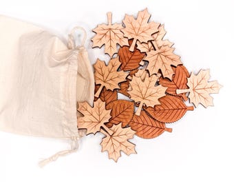 May include: A collection of laser-cut wooden leaf shapes in light and medium brown tones, featuring maple and rounded leaf designs with etched vein details, spilling out from an open, off-white cotton drawstring storage pouch.