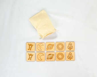 May include: A wooden space-themed memory game with a beige drawstring bag. The game features eight wooden square tiles with engraved images of an astronaut, moon, planet, sun, and rocket. The tiles are laid out on a white surface.
