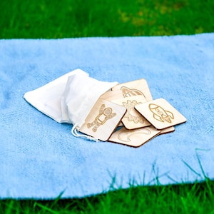 Ten-piece natural colored wooden space memory match game with astronaut, rocket, and sun tiles spilling from beige cotton bag onto blue Montessori floor mat in grass.