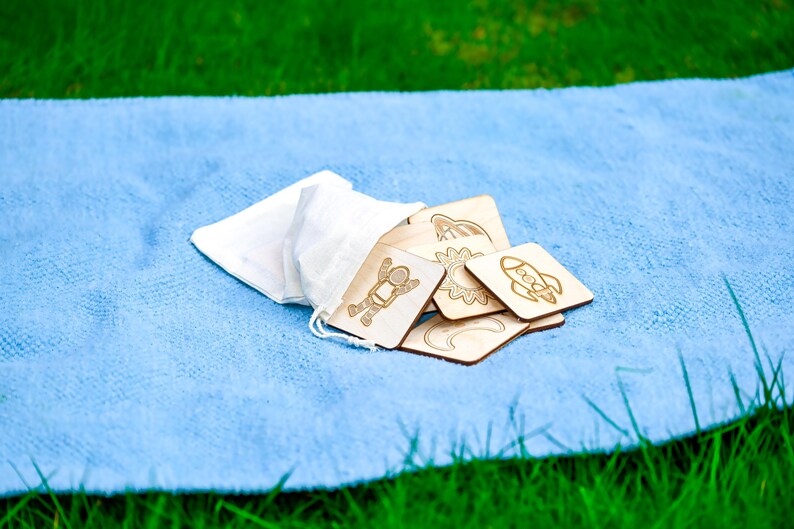 Ten-piece natural colored wooden space memory match game with astronaut, rocket, and sun tiles spilling from beige cotton bag onto blue Montessori floor mat in grass.