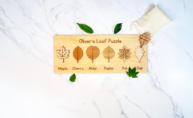 Personalized Montessori leaf puzzle engraved 'Oliver's Leaf Puzzle' with six labeled wooden leaves - maple, cherry, alder, poplar, ash, and oak - the oak piece resting on a muslin bag beside fresh green leaves on marble.