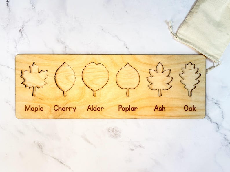Empty leaf puzzle board showing six leaf-shaped recesses with engraved name labels - maple, cherry, alder, poplar, ash, oak - in neutral Baltic birch beside a muslin bag.