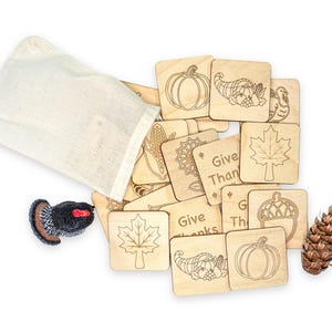 May include: A set of square wooden tiles featuring engraved autumn-themed illustrations including pumpkins, corn, leaves, acorns, and cornucopias, alongside text reading Give Thanks, accompanied by a fabric drawstring bag, a turkey figurine, and a pinecone.