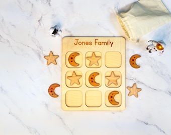 May include: A wooden tic-tac-toe board with the text Jones Family engraved at the top. The game includes wooden star and crescent moon pieces, two small astronaut figurines, and a beige fabric drawstring storage bag for the game pieces.