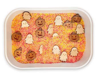 May include: A white rectangular plastic bin filled with a mixture of yellow, orange, and pink dyed rice. Scattered throughout the rice are several small, flat, wooden cutouts shaped like smiling ghosts and jack-o'-lantern pumpkins.