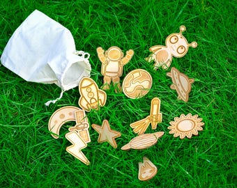 May include: Wooden space-themed cutouts, such as an astronaut, rockets, planets, and an alien, are scattered on green grass beside a white drawstring bag. The cutouts are light brown with etched details.