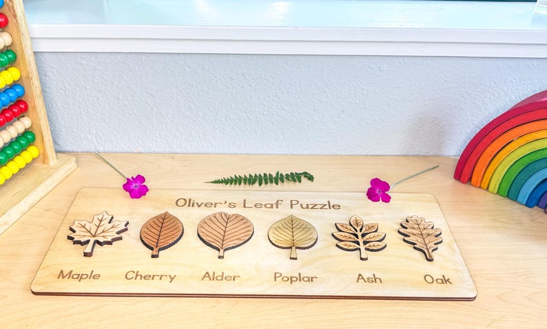 Personalized leaf puzzle on a playroom desk with a fern frond, pink blossoms, wooden rainbow stacker, and abacus, six labeled wooden leaves seated in the board. The wooden leaves are labeled - maple, cherry, alder, poplar, ash, and oak.  Each leaf is laser-engraved with realistic veins above its engraved name label.
