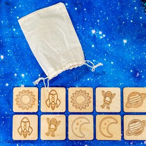 Ten-piece neutral-colored wooden space memory match set arranged on starry night backdrop, featuring five pairs of laser-engraved astronaut, moon, planet, sun, rocket tiles.