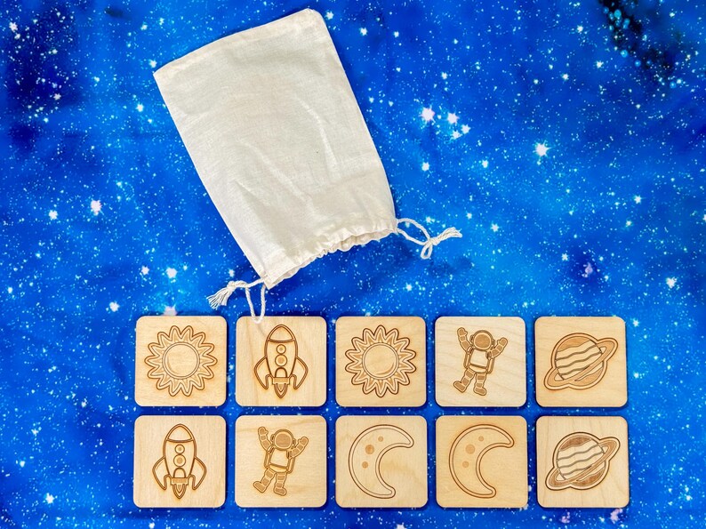 Ten-piece neutral-colored wooden space memory match set arranged on starry night backdrop, featuring five pairs of laser-engraved astronaut, moon, planet, sun, rocket tiles.