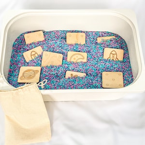 10 wooden space memory match tiles hidden in white sensory bin filled with blue, pink, and purple rice. Shown with natural cotton drawstring storage pouch on a white fabric background.