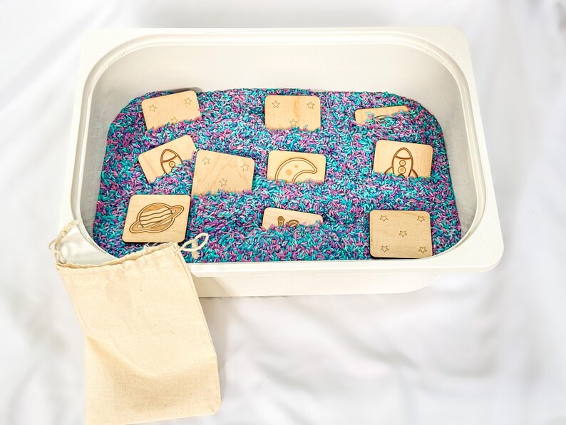 10 wooden space memory match tiles hidden in white sensory bin filled with blue, pink, and purple rice. Shown with natural cotton drawstring storage pouch on a white fabric background.