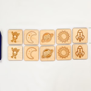 Ten-piece neutral colored wooden space memory match game shown beside jumbo crayon for size reference, featuring astronaut, moon, Saturn, sun, rocket pairs.  On a light white fabric background.
