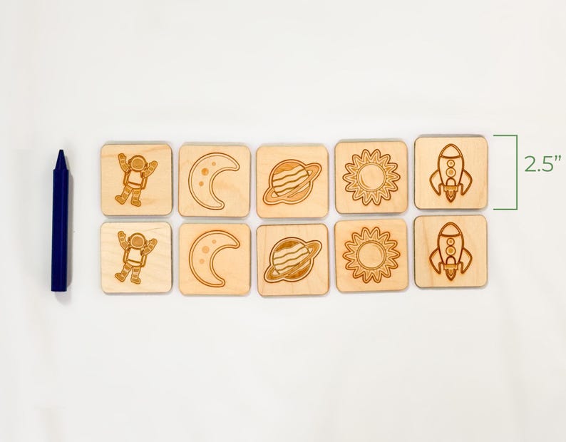 Ten-piece neutral colored wooden space memory match game shown beside jumbo crayon for size reference, featuring astronaut, moon, Saturn, sun, rocket pairs.  On a light white fabric background.