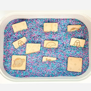 Ten double-sided natural colored wooden space memory tiles partially buried in purple and teal rainbow rice sensory bin.