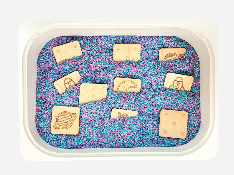 Ten double-sided natural colored wooden space memory tiles partially buried in purple and teal rainbow rice sensory bin.