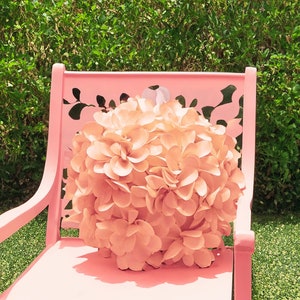 May include: A pink metal chair with a floral cutout design. A large, peach-colored flower sits on the seat of the chair.