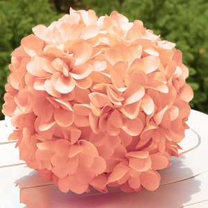 May include: A peach-colored paper flower ball, made of many individual paper flower petals, is sitting on a white surface.