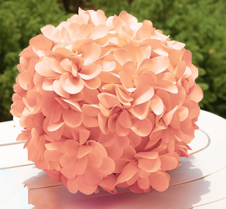 May include: A peach-colored paper flower ball, made of many individual paper flower petals, is sitting on a white surface.