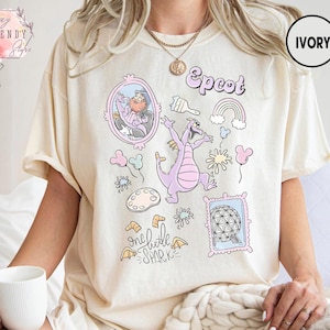 May include: Ivory-colored t-shirt featuring a whimsical design with the word "Epcot" in a playful font. The design includes a purple dragon, a rainbow, and other cartoon elements. The shirt is a relaxed fit.