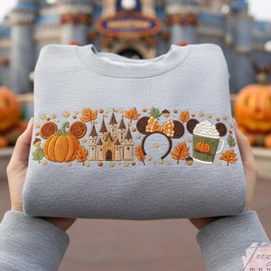 May include: A light grey crewneck sweatshirt featuring an embroidered horizontal band of autumn-themed icons, including a pumpkin, a castle, mouse ears with a bow, and a coffee cup with whipped cream, surrounded by orange and brown fall leaves.