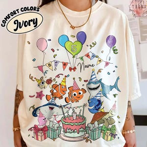 May include: Ivory-colored Comfort Colors t-shirt featuring a colorful birthday design. The design includes balloons, a banner, and cartoon characters. The number 5 is displayed on a balloon, and a space is available for a name.