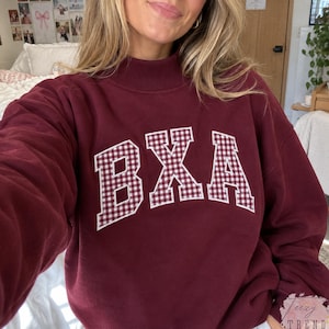 May include: A maroon long-sleeve sweatshirt featuring a mock neck collar and the letters BXA across the chest. The letters are filled with a white and maroon gingham check pattern and outlined in white, creating a bold, collegiate-style graphic.