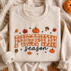 Könnte beinhalten: Ein cremefarbenes Sweatshirt mit Rundhalsausschnitt und herbstlichen Stickereien. Der Schriftzug PUMPKIN besteht aus orange-weiß karierten Blockbuchstaben, ergänzt durch das Wort season in schwarzer Schreibschrift, umgeben von Kürbissen und Blättern.