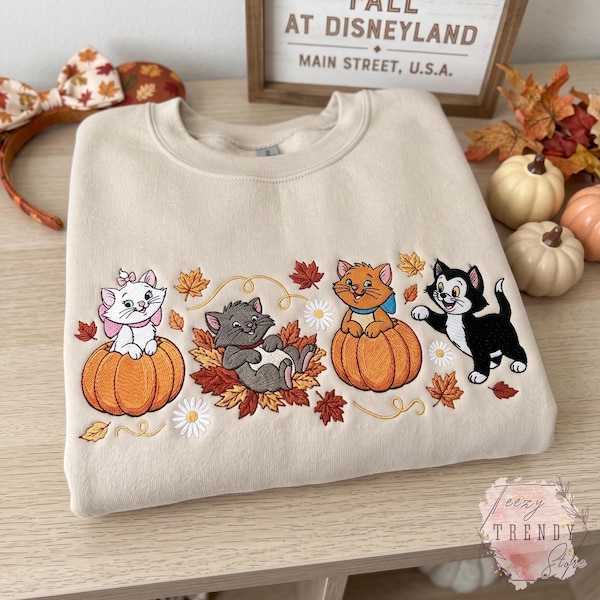 May include: A beige crewneck sweatshirt featuring embroidered Disney Aristocats characters including Marie, Berlioz, and Toulouse. The design includes orange pumpkins, autumn leaves, and white daisies. The sweatshirt is displayed with a pair of fall-themed ears.