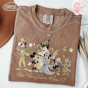 Peut inclure: T-shirt couleur espresso avec un motif de personnages Disney célébrant le Nouvel An. Le motif comprend Mickey et Minnie Mouse, Donald et Daisy Duck et Dingo. Le texte "New Year's Eve on Main St." est imprimé en dessous. Un collier argenté est posé sur le t-shirt.