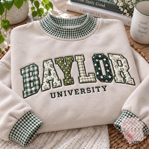 May include: A beige crewneck sweatshirt featuring a green and white gingham collar and cuffs. The word BAYLOR is appliqued in a mix of green gingham, floral, and star patterns, with the word UNIVERSITY embroidered in dark green below.
