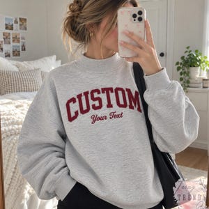 May include: A light gray crewneck sweatshirt featuring the word CUSTOM in large, arched, dark red capital letters, with the words Your Text written in a smaller, cursive dark red font directly underneath the main text on the chest.