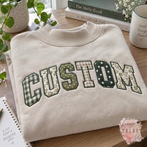 Könnte beinhalten: Ein beigefarbenes Sweatshirt mit Rundhalsausschnitt und dem Wort CUSTOM in großen, applizierten Blockbuchstaben. Jeder Buchstabe besteht aus verschiedenen Stoffen, darunter grünes Vichy-Karo, florale Muster, olivfarbene Punkte und Dunkelgrün mit weißen Sternen.