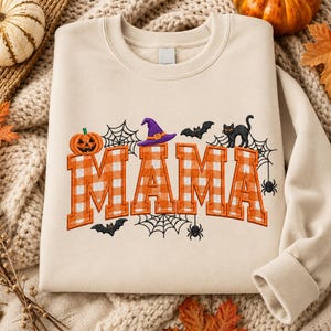 Könnte beinhalten: Ein beiges Sweatshirt mit Rundhalsausschnitt und dem gestickten Wort MAMA in orange-weißem Vichy-Muster. Das Design zeigt einen Kürbis, einen violetten Hexenhut, schwarze Fledermäuse, eine schwarze Katze sowie Spinnennetze mit kleinen Spinnen.