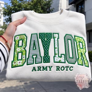 May include: A cream-colored crewneck sweatshirt featuring the word BAYLOR in large, green-bordered block letters, each filled with different green and white patterns, and the text ARMY ROTC embroidered in green below the main design.