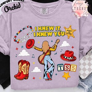 May include: A light purple t-shirt featuring a graphic of a person with long hair playing a guitar, surrounded by cowboy boots, hats, a beaded bracelet with the text 'you've got a friend in me', a sheriff badge, and the text 'I KNEW IT, I KNEW YOU'.
