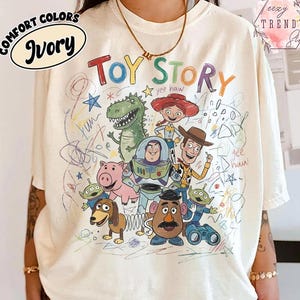 May include: Ivory-colored t-shirt featuring a colorful graphic of Toy Story characters. The design includes Woody, Buzz Lightyear, and other characters from the animated film. The words "Toy Story" are written in a playful font.