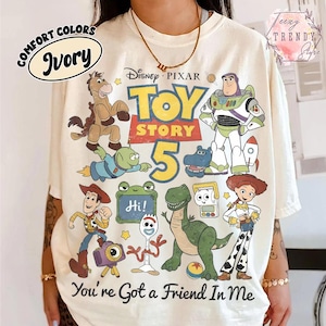 May include: An ivory t-shirt featuring a graphic print of Disney Pixar Toy Story 5 characters including Woody, Buzz Lightyear, Jessie, Rex, Bullseye, and Forky. The text reads Toy Story 5 and You're Got a Friend In Me with a small frog sign saying Hi.