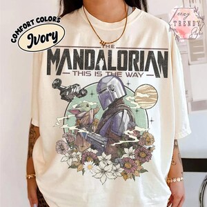 May include: Ivory t-shirt with a graphic of The Mandalorian and Grogu surrounded by flowers. The text "THE MANDALORIAN - THIS IS THE WAY" is printed above the image. The shirt has the Comfort Colors brand logo.