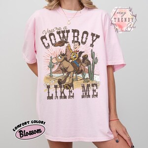 May include: A light pink short-sleeve t-shirt featuring a graphic of a cartoon cowboy riding a horse in a desert landscape with cacti. The text reads You're a COWBOY LIKE ME. The design includes a sunburst background and small flowers.