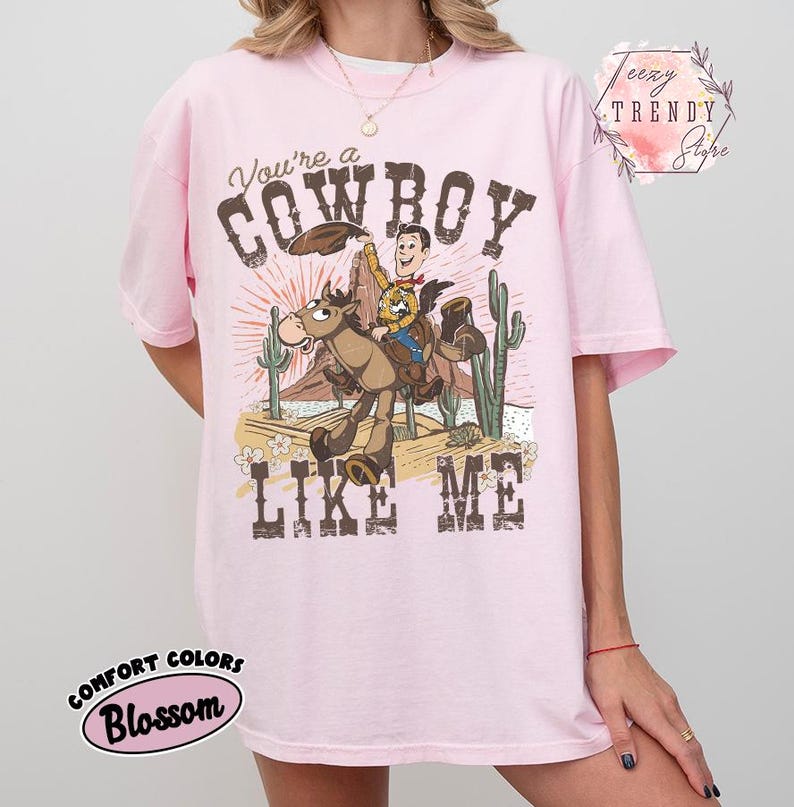 May include: A light pink short-sleeve t-shirt featuring a graphic of a cartoon cowboy riding a horse in a desert landscape with cacti. The text reads You're a COWBOY LIKE ME. The design includes a sunburst background and small flowers.