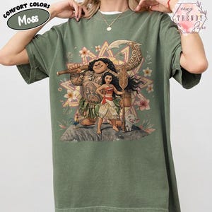 Pu&ograve; includere: T-shirt verde muschio con una grafica colorata di personaggi dei cartoni animati. Il design include una grande figura tatuata, una giovane donna e un maialino. La maglietta presenta le scritte "Comfort Colors" e "Moss".