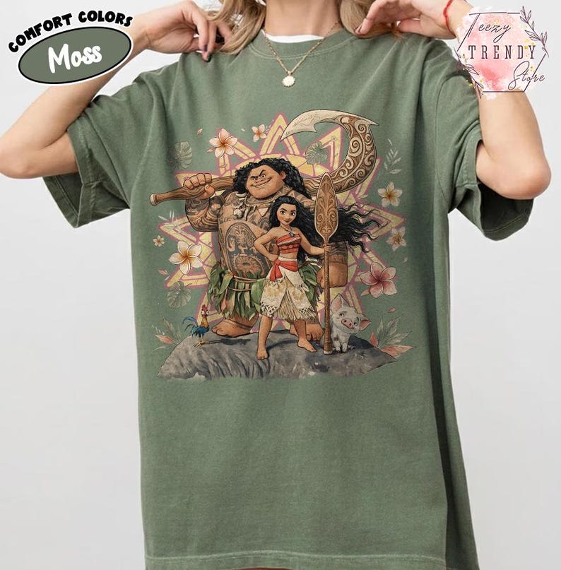 Pu&ograve; includere: T-shirt verde muschio con una grafica colorata di personaggi dei cartoni animati. Il design include una grande figura tatuata, una giovane donna e un maialino. La maglietta presenta le scritte "Comfort Colors" e "Moss".