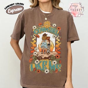 May include: An espresso brown short-sleeve t-shirt featuring a graphic print of two figures in cowboy hats standing together. The design includes the text COWGIRL LIKE ME, a sun, flowers, and flames in shades of orange, yellow, blue, and white.