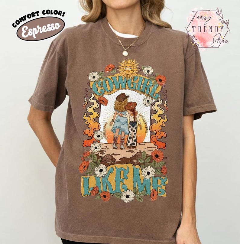May include: An espresso brown short-sleeve t-shirt featuring a graphic print of two figures in cowboy hats standing together. The design includes the text COWGIRL LIKE ME, a sun, flowers, and flames in shades of orange, yellow, blue, and white.