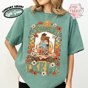May include: A light green short-sleeve t-shirt featuring a graphic print of two figures in cowboy hats with the text "COWGIRL LIKE ME". The design includes a sun, orange flames, and white and red flowers surrounding the central illustration.