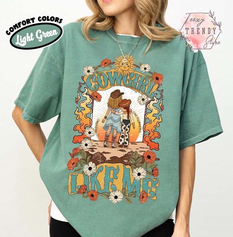 May include: A light green short-sleeve t-shirt featuring a graphic print of two figures in cowboy hats with the text "COWGIRL LIKE ME". The design includes a sun, orange flames, and white and red flowers surrounding the central illustration.